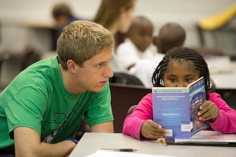 A student helps a young girl read a book.