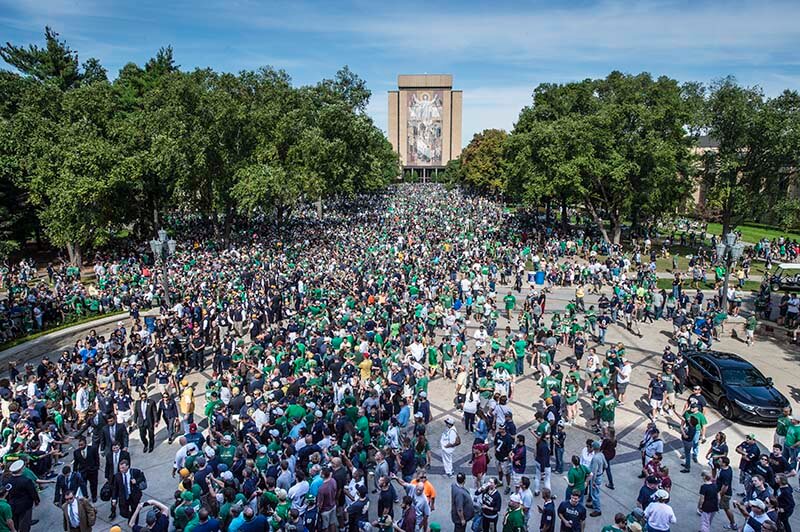 Fans wearing green gather in the Quad, with Touchdown Jesus in the background.